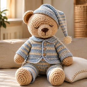 Puede incluir: Un oso de peluche tejido a ganchillo con hilo color canela, que lleva un pijama de rayas azules y blancas con tres pequeños botones de madera y un gorro de dormir a juego con un pequeño pompón. El oso tiene los ojos cerrados y nariz marrón.