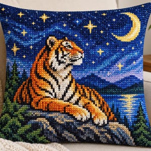 May include: A square throw pillow featuring a cross-stitch design of an orange and black tiger resting on a rocky cliff. The background depicts a dark blue night sky with a yellow crescent moon, twinkling stars, distant mountains, and pine trees.
