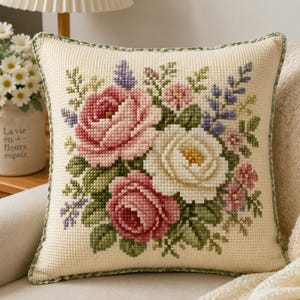 May include: A square throw cushion featuring a cross-stitch floral pattern on a cream-coloured fabric. The design displays a central bouquet of pink and white roses, accented with small purple flowers and green leaves, finished with a green piped edge.