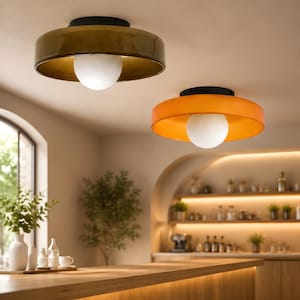 May include: Two modern ceiling lights with a round, flat design. One is olive green, the other is orange, both with a black top and a white globe light. The lights are in a kitchen setting with a wooden countertop and a window.