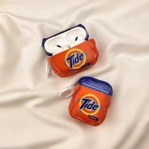 Pode incluir: Duas capas protetoras laranja e azul para fones de ouvido sem fio, projetadas para se parecerem com cápsulas de detergente Tide. Uma capa está aberta, revelando fones brancos dentro, enquanto a outra está fechada. Ambas exibem o logotipo Tide.