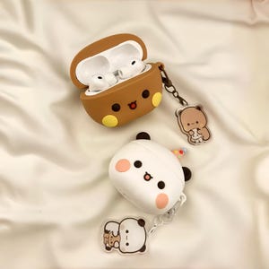 Puede incluir: Dos fundas de silicona para auriculares con caras de personajes. Una funda marrón está abierta, mostrando auriculares blancos, con un dije de oso. La otra es una funda blanca cerrada con un pequeño gorro de fiesta y un dije de panda.