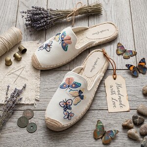 May include: A pair of off-white canvas espadrilles featuring embroidered colorful butterflies on the upper. The insoles are printed with the text A couple of lines. A tan paper tag attached to the shoe reads Handmade with Love in black script.