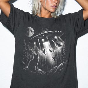 Könnte beinhalten: Ein anthrazitfarbenes Kurzarm-T-Shirt mit einem Grafikdruck eines UFOs, das über einem Wald schwebt und Lichtstrahlen auf mehrere Katzen wirft. Ein Vollmond ist oben links zu sehen. Das Wort GRAPHITE ist unten links aufgedruckt.