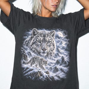 K&ouml;nnte beinhalten: Ein anthrazitfarbenes Kurzarm-T-Shirt mit einem detaillierten Grafikdruck, der das Gesicht eines Schneeleoparden und einen kleineren Schneeleoparden zeigt, der unter einem Vollmond durch eine verschneite Berglandschaft l&auml;uft. Das Wort GRAPHITE ist aufgedruckt.