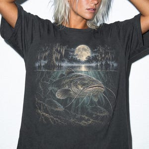 Op de afbeelding: Een antracietgrijs T-shirt met korte mouwen en een grafische print van een grote meerval die onder water zwemt in een moerassige omgeving met cipressen en een volle maan op de achtergrond. Het woord GRAPHITE staat in de linkerbenedenhoek.