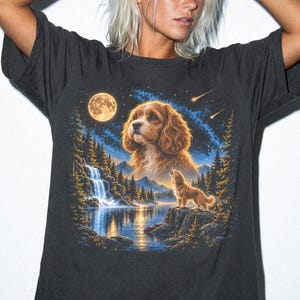 May include: A charcoal gray t-shirt featuring a graphic print of a brown spaniel dog, a smaller dog howling on a rock, a waterfall, a lake, a full moon, and a starry night sky. The word GRAPHITE is printed in black in the bottom left corner.