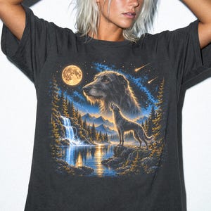 May include: A charcoal grey short-sleeve t-shirt featuring a graphic print of a wolf howling at a full moon above a mountain lake and waterfall. The design includes a large wolf head in the starry night sky and a smaller wolf standing on a rock.