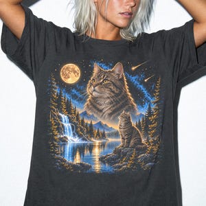 Könnte beinhalten: Ein anthrazitfarbenes T-Shirt mit einem Grafikdruck einer getigerten Katze, die in einem sternenklaren Nachthimmel auf einen Vollmond blickt. Die Szene zeigt einen Wasserfall, einen See und eine kleine Katze auf einem Felsen. Das Wort GRAPHITE ist aufgedruckt.