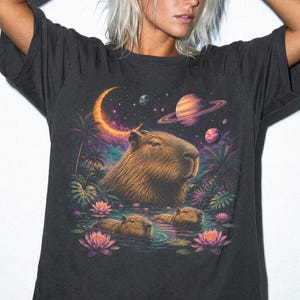 May include: A charcoal gray t-shirt featuring a graphic print of a large capybara head above two smaller capybaras swimming in a pond with pink lotus flowers, palm trees, a crescent moon, and planets in a colorful, vibrant space-themed sky.