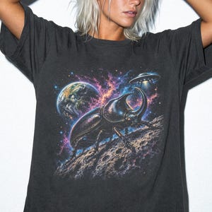 May include: A charcoal gray short-sleeve t-shirt featuring a graphic print of a large rhinoceros beetle on a cratered surface in space. The design includes a colorful nebula, the planet Earth, and a flying saucer hovering in the dark starry background.