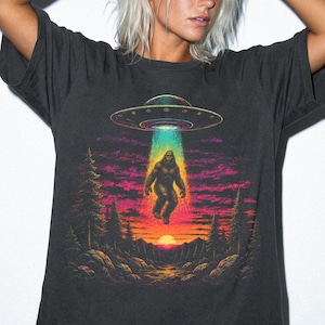 Könnte beinhalten: Ein graphitfarbenes T-Shirt mit einem Grafikdruck, der eine große, dunkle, haarige Kreatur zeigt, die von einer fliegenden Untertasse entführt wird. Die Szene umfasst einen Sonnenuntergang, silhouettierte Kiefern und einen leuchtenden Lichtstrahl.