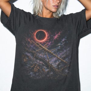 May include: A charcoal gray short-sleeve t-shirt featuring a graphic print of a large stick insect resting on a branch under a solar eclipse in a dark, starry sky with a faint flying saucer visible in the background. The word GRAPHITE is printed below.