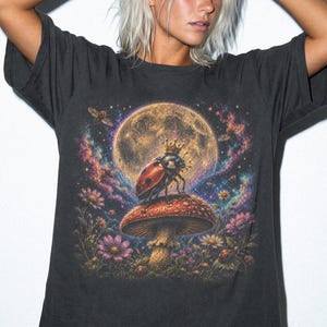Puede incluir: Una camiseta gris carbón de manga corta con un estampado gráfico de una mariquita con corona, posada sobre una seta grande. El fondo muestra una luna llena rodeada de coloridas nubes de nebulosa, pequeñas flores y follaje.