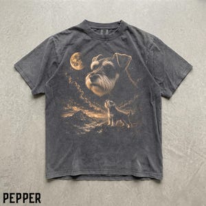 Könnte beinhalten: Ein anthrazitfarbenes Kurzarm-T-Shirt mit einem Grafikdruck eines Schnauzers, der zu einem großen Mond über einer Berglandschaft aufblickt, während ein kleinerer Schnauzer auf einem Felsgipfel unter einem Sternenhimmel steht.