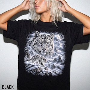 K&ouml;nnte beinhalten: Ein schwarzes T-Shirt mit kurzen &Auml;rmeln und einem gro&szlig;en Grafikdruck eines Schneeleoparden. Das Design zeigt das Gesicht des Leoparden in einer Nahaufnahme &uuml;ber einem kleineren, ganz abgebildeten Leoparden in einer verschneiten Berglandschaft.