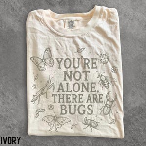 Peut inclure: Un t-shirt ivoire avec le texte gris foncé YOU'RE NOT ALONE, THERE ARE BUGS imprimé au centre. Le motif comprend des illustrations au trait détaillées de divers insectes, notamment des papillons, une mante religieuse, un scarabée et une abeille.