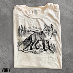 以下が含まれることがあります： アイボリー色のTシャツで、岩の上に立つキツネの黒いインクスタイルのグラフィックが描かれています。背景には湖、松の木、山々が描かれています。首元の内側のラベルには「Comfort Colors, 100% Cotton, Made in Honduras」という文字が記されています。