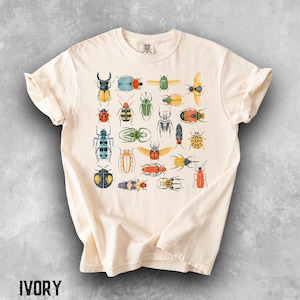 以下が含まれることがあります： アイボリー色の半袖Tシャツで、中央に様々なカラフルな甲虫や昆虫のグラフィックプリントが施されています。赤、黄、緑、青、オレンジの色合いで描かれた多種多様な昆虫のコレクションが、グリッド状のパターンで配置されています。