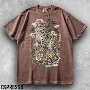 以下が含まれることがあります： 茶色の半袖Tシャツ。岩の上に座るユキヒョウと、その周りに描かれた花、松の木、雲のグラフィックプリントが特徴。ヒョウの右側には、縦長の長方形の枠の中に日本語のテキストがデザインされている。