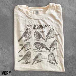 May include: An ivory t-shirt featuring a dark brown graphic print titled North American Sparrows. The design displays detailed illustrations of eight different sparrow species, each labeled with its common name, including Song, Chipping, and Fox.