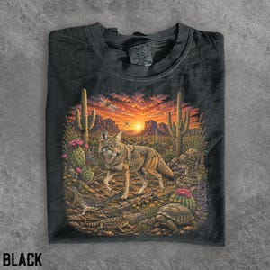 Peut inclure: Un t-shirt noir avec une impression graphique représentant un coyote dans un paysage désertique au coucher du soleil. La scène inclut des cactus saguaro, un grand géocoucou, une tortue et un lézard. Étiquette Comfort Colors au col.