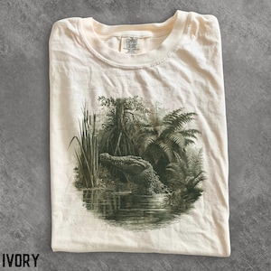 Może przedstawiać: T-shirt z krótkim rękawem w kolorze kości słoniowej ze szczegółowym ciemnozielonym nadrukiem graficznym przedstawiającym aligatora wyłaniającego się z bagiennej wody w otoczeniu pałek wodnych, paproci i namorzynów. Wewnętrzna metka zawiera napis Comfort Colors.
