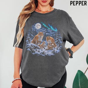 Puede incluir: Una camiseta de manga corta gris carbón con un estampado gráfico de tres zorros en un paisaje forestal nevado bajo una luna llena y la aurora boreal. El texto PEPPER está impreso en la esquina superior derecha de la imagen.