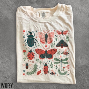 May include: An ivory t-shirt featuring a graphic print of various insects including beetles, butterflies, moths, dragonflies, bees, and caterpillars in shades of red, dark green, and black, interspersed with small floral and star motifs.