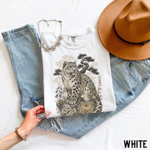 以下が含まれることがあります： 岩の上に座るユキヒョウの緻密なイラストが描かれた白いTシャツ。松の木と花が周囲に配置されています。このTシャツは、ライトブルーのデニムジーンズ、茶色のフェドラハット、シルバー調のフラワーネックレスと組み合わされています。
