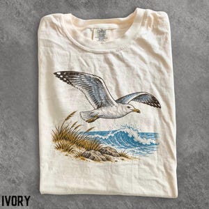 Op de afbeelding: Een ivoorkleurig t-shirt met ronde hals en een grafische print van een vliegende meeuw boven een kustlandschap met blauwe oceaangolven, beige helmgras en grijze rotsen. Het shirt heeft een Comfort Colors label in de binnenhals.
