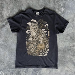 以下が含まれることがあります： チャコールグレーの半袖Tシャツ。雪豹が様式化された花、雲、松の木の中に座っている詳細なグラフィックプリントが施されています。デザインの右側には、小さな長方形の中に縦書きの日本語のテキストが含まれています。
