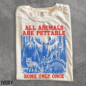 以下が含まれることがあります： アイボリーのTシャツで、赤色のグラフィックプリントには「ALL ANIMALS ARE PETTABLE SOME ONLY ONCE」と書かれています。テキストの下には、森の中でクマ、ヘラジカ、オオカミ、キツネ、アライグマ、ヤマアラシが描かれた青いイラストが配置されています。