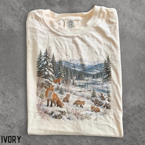 Peut inclure: Un t-shirt à col rond couleur ivoire arborant une impression représentant un paysage hivernal enneigé avec une rivière, des sapins et des montagnes. L'image montre cinq renards roux de tailles différentes sur le terrain enneigé.