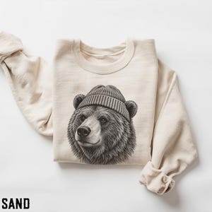 Op de afbeelding: Een zandkleurige sweater met ronde hals en een gedetailleerde zwart-witillustratie van een beer met een geribbelde muts. Het kledingstuk heeft lange mouwen en een ronde hals, met het woord SAND in het zwart in de linkerbenedenhoek gedrukt.