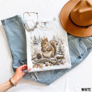 May include: A white t-shirt featuring a graphic of a squirrel holding an acorn in a snowy forest setting with pine trees and pinecones. The shirt is paired with blue denim jeans, a tan fedora hat, and a silver-toned floral necklace.