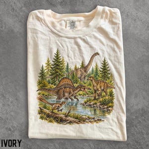 Op de afbeelding: Een ivoorkleurig t-shirt met een grafische print van diverse dinosaurussen in een bosrijke omgeving bij een waterpartij. De afbeelding toont een langnekkige sauropode, een dinosaurus met rugkam en een kleine theropode bij een boomstam.
