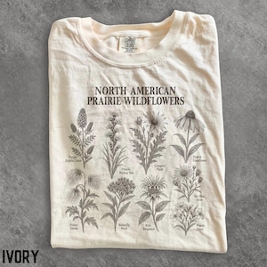 May include: An ivory t-shirt featuring the text North American Prairie Wildflowers and detailed black line drawings of various plants including Purple Prairie Clover, Prairie Blazing Star, Compass Plant, Prairie Coneflower, Prairie Smoke, Butterfly Weed, Wild Bergamot, Sky Blue Aster, and Prairie Phlox.