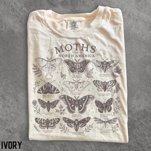 Op de afbeelding: Een crèmekleurig t-shirt met een donkerbruine grafische print met de titel MOTHS OF NORTH AMERICA. Het ontwerp toont twaalf gedetailleerde illustraties van verschillende mottensoorten met hun wetenschappelijke namen in kleine letters eronder.