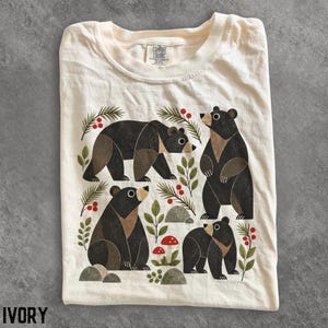 Pode incluir: Uma t-shirt cor de marfim com uma impressão gráfica de quatro ursos pretos em várias poses, rodeados por ramos de pinheiro verdes, bagas vermelhas, pequenas rochas cinzentas e dois cogumelos vermelhos e brancos. Etiqueta Comfort Colors.