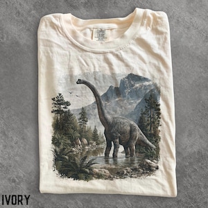 Op de afbeelding: Een ivoorkleurig t-shirt met een grafische print van een dinosaurus met een lange nek die in een ondiepe rivier staat, omringd door dennenbomen en bergen. Het shirt heeft een ronde hals en een Comfort Colors label in de binnenkraag.