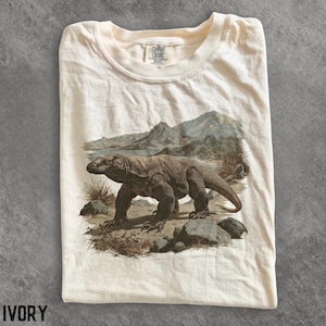 Può includere: Una t-shirt color avorio con una stampa grafica di un drago di Komodo che cammina su un terreno roccioso con montagne sullo sfondo. La maglietta presenta un girocollo e un'etichetta del marchio Comfort Colors stampata all'interno del colletto.