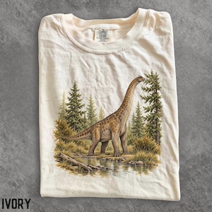 May include: An ivory-coloured t-shirt featuring a graphic print of a long-necked sauropod dinosaur standing in a forest landscape with pine trees, tall grasses, and a small stream. The shirt has a crew neckline and a Comfort Colors brand label.