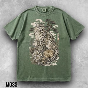 以下が含まれることがあります： モスグリーンの半袖Tシャツ。岩の上に座るユキヒョウのグラフィックが描かれており、周囲には様式化された花、雲、松の木が配置されている。グラフィックには、縦長の長方形の枠内に日本語の文字が含まれている。