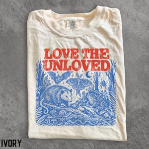 May include: An ivory t-shirt featuring the red text LOVE THE UNLOVED above a blue graphic illustration of a forest scene. The design includes an opossum, a moth, a bat, a toad, a snail, a beetle, a snake, a spider, and a crescent moon.