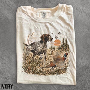 May include: An ivory t-shirt featuring a graphic print of a brown and white spotted hunting dog standing in tall grass near a pheasant in flight. The scene includes a sunset, pine trees, and birds in the sky. The shirt has a Comfort Colors label.