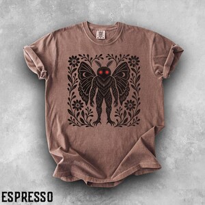 May include: An espresso-colored short-sleeve t-shirt featuring a black graphic print of a moth-like creature with glowing red eyes. The creature is centered within a square frame of black floral vines and leaves. The shirt has a crew neckline.