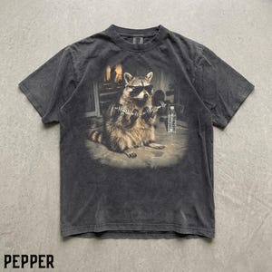 May include: A charcoal grey short-sleeve t-shirt featuring a graphic print of a raccoon wearing black sunglasses and lifting dumbbells. The raccoon is positioned in a gym setting with a water bottle nearby. The shirt has a faded, vintage-style wash.