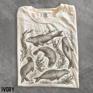 以下が含まれることがあります： アイボリー色のTシャツで、海藻や泡の間を泳ぐ4頭のあざらしが黒いインクで詳細に描かれています。Tシャツはクルーネックで、襟の内側にComfort Colorsのブランドラベルが付いています。あざらしは様々なポーズで描かれています。