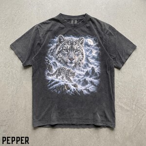 K&ouml;nnte beinhalten: Ein anthrazitfarbenes Kurzarm-T-Shirt mit einem Grafikdruck eines Schneeleopardengesichts und einem kleineren Schneeleoparden, der unter einem Vollmond &uuml;ber ein verschneites Gebirge l&auml;uft. Das Shirt hat einen Rundhalsausschnitt und eine verwaschene Optik.